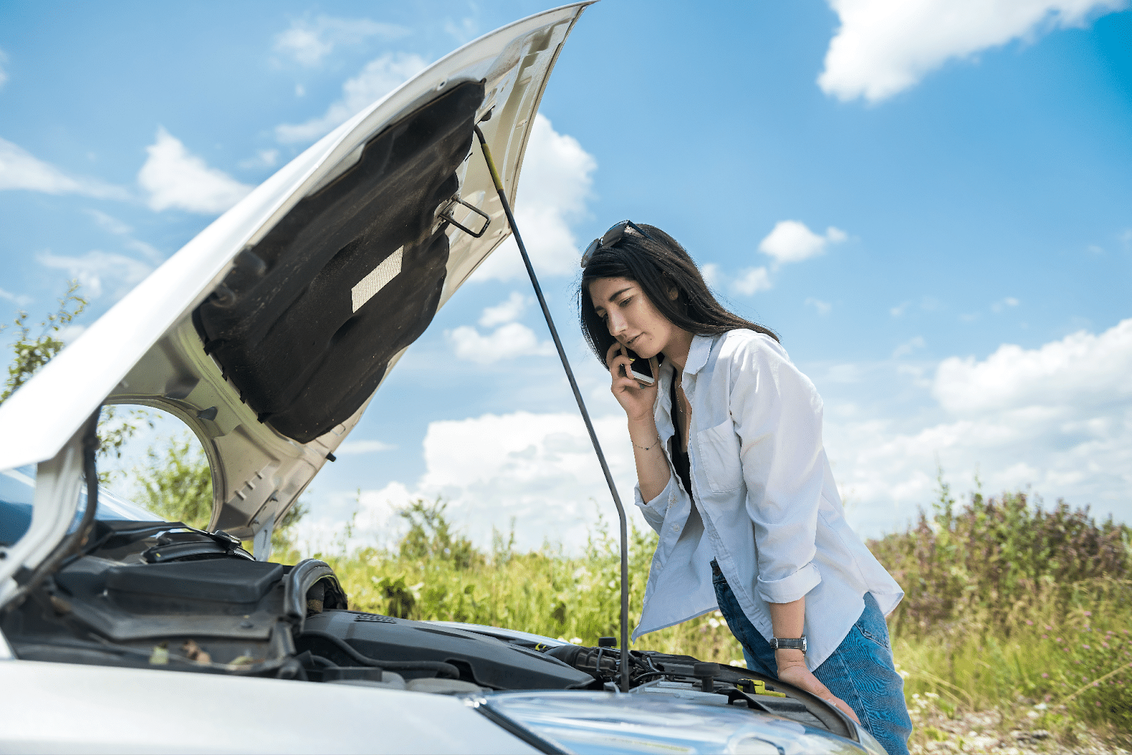Young woman looks under the car bonnet due to a car breakdown in summer.